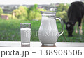 A glass and jug filled with fresh milk are placed on a wooden surface, set against a lush green field and a grazing cow. The scene highlights organic farming and healthy nutrition 138908506
