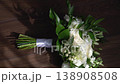 A sophisticated wedding bouquet featuring fresh white roses, lush greenery, and small white berries rests on polished wood. Ideal for modern bridal themes and romantic floral arrangements 138908508