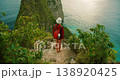 Woman with backpack looks at steep green cliff and blue ocean at Kelingking Nusa Penida Bali 138920425