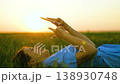 Gadget in hands of younger generation. Girl lies on green grass uses smartphone. Happy teenager smiles with gadget, green lawn, sunset, park. Technology for people, outdoor recreation. People gadgets 138930748
