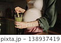 Pregnant woman with a big belly standing in a sunlit kitchen, stirring and drinking a green detox smoothie from a glass, promoting a healthy lifestyle and proper nutrition during pregnancy 138937419