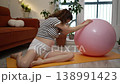 Expectant woman performing prenatal yoga exercises at home, using a large pink fitness ball on a yellow mat to stretch her back and hips, actively preparing her body for childbirth 138991423