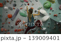 Strong girl in sports outfit is climbing up colorful artificial wall in gym gripping rocks focused of extreme activity. Youth, entertainment and training concept. 139009401