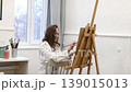 Young female artist standing near easel and holding pencil while working on painting in art studio, creative process and inspiration 139015013