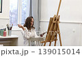 Young female artist standing near easel and holding pencil while working on painting in art studio, creative process and inspiration 139015014