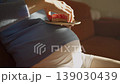Pregnant woman resting on a cozy couch satisfies her sweet cravings by eating a pink-glazed donut from a small plate placed on her large, round belly, basking in warm sunlight 139030439