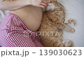 Top-down view of a pregnant woman in pajamas tenderly stroking her bare belly while her fluffy ginger cat rests contentedly beside her, showing a heartwarming bond of affection 139030623