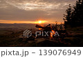 A blazing bonfire atop a mountain in the Karkonosze range, overlooking the setting sun during the golden hour, with Sniezka peak visible in the background. 139076949