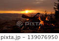A blazing bonfire atop a mountain in the Karkonosze range, overlooking the setting sun during the golden hour, with Sniezka peak visible in the background. 139076950