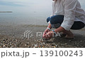 A woman squats barefoot on a serene pebble beach in Malaga, gently collecting seashells and small stones at the water's edge. The calm sea and rhythmic waves create a tranquil, meditative atmosphere. 139100243