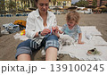 A caring mother assists her young son with his shoes on a peaceful beach in Malaga, capturing a heartwarming family vacation moment. 139100245