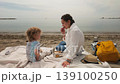 A mother and her young child share a tender moment during a picnic on the sandy beach. They enjoy their time together, fostering a loving bond by the peaceful ocean. 139100250