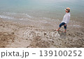 A serene woman in a hat enjoys a peaceful walk barefoot along the tranquil sandy beach. Gentle sea waves wash over her feet, creating a calming and relaxing vacation moment. 139100252