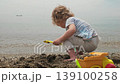 An adorable young child enjoys a peaceful day at the beach, happily playing with sand and toys by the gentle ocean waves. 139100258