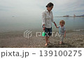 A heartwarming scene of a mother and her young son walking barefoot on the sandy beach, holding hands and carrying a small bucket. They share a tender moment by the sea. 139100275