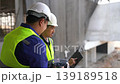 Two Asian construction engineers or site supervisors wearing hard hats and safety vests discussing project details while reviewing digital blueprints on a laptop and using a walkie-talkie at a buildin 139189518