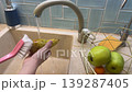In a bright home kitchen, a vegan kid carefully scrubs dirt from freshly picked pear under clean running water, learning good hygiene. 139287405