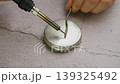 Detailed shot of a person using a soldering iron to melt solder wire onto a round metal jar lid on a grey workbench surface 139325492