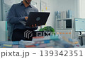 Thoughtful young Latin businessman stand with laptop analyzing data above paperwork stack. Hybrid work audit, digital transformation strategy, business corporate finance, executive decision concept. 139342351
