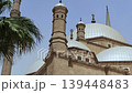Low angle view of Muhammad Ali Mosque domes, minarets and crescent finials at Cairo Citadel, Egypt, with palm tree framing Ottoman Islamic architecture against hazy purple sky. 139448483