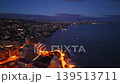 Drone Dusk Coastline City Lights Glow Over Bay, Illuminated Waterfront Roads, Waves Lapping Cliffs, Distant 139513711