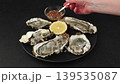 Khasan oysters, live, fresh, opened. Served with classic French Mignonette sauce and half a lemon. The woman adds sauce. Blue plate, black background. Selective focus, close-up. 139535087