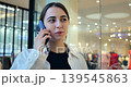 Smiling woman engaging on mobile near store. Portrait of joyful woman talking on phone beside shop windows. Cheerful woman with friendly expression communicating on phone near retail storefronts 139545863