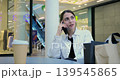 Young woman seated at mall cafe table during phone call, resting between purchases with coffee cup and shopping bags, checking order details, relaxed attentive expression, escalator and modern 139545865