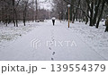 A lone person walks along a snow-covered path in a quiet pine park during a snowfall. Footprints mark the trail as the silhouette strolls toward the hazy, mist-filled forest. Peaceful winter scene 139554379