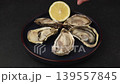 Moari oysters, live, fresh, opened. Served with half a lemon. A woman pours lemon juice over an oyster. Blue plate, black background. Selective focus, close-up. 139557845