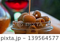 Hand lifts olive appetizer on skewer above glass jar during outdoor snack scene. Wine glass appears in background, warm light and shallow depth of field create appetizing Mediterranean mood. 139564577