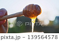 Hand holds wooden spoon with golden honey dripping into container in warm backlit outdoor scene. Sunlight makes liquid glow with rich amber color and natural texture. 139564578