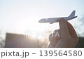Person raises miniature airplane in front of sun, as if guiding it through air above rural setting. Scene suggests travel, imagination, and playful motion in natural light. 139564580