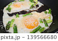 Cooking process of eggs and asparagus in a frying pan, showcasing the gradual melting of cheese and bubbling of egg whites in a kitchen setting 139566680
