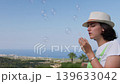 Calm Woman Blowing Bubbles in Sunlight, Sky and Sea Background 139633042