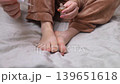 A woman in pajamas sitting on a bed and applying antifungal nail polish to treat onychomycosis. Home treatment of fungal infection, yellow nails, and medical pedicure for healthy feet. A woman in pajamas sitting on a bed and applying antifungal nail polish to treat onychomycosis. Home treatment of fungal infection, yellow nails, and medical pedicure for healthy feet. 139651618