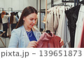 Smiling young woman holding pink dress on hanger in clothing store, carefully choosing stylish outfit during shopping day 139651873