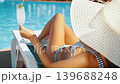 Woman in bikini and straw hat relaxing on sunbed by swimming pool. Female holding refreshing cocktail and enjoying summer luxury resort 139688248