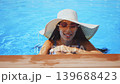 Smiling young woman in white straw hat and sunglasses relaxing in blue swimming pool. Female enjoying summer vacation in luxury resort water 139688423
