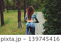Young woman riding bicycle along narrow paved path in green forest, seen from behind, wearing denim outfit, enjoying outdoor activity 139745416