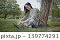 A hand-held shot of a young female student in casual clothes wearing glasses sitting on the grass under a tree in a city park and using a laptop typing on a keyboard. 139774291