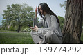 The shot is handheld and shows a young female student in casual clothes wearing glasses sitting on the grass under a tree in a park and using a mobile phone. 139774292