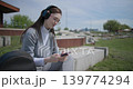 A hand-held shot of a young female student in casual clothes, wearing glasses and headphones, sitting on a concrete in an urban park on a sunny summer day and listening to music from a mobile phone 139774294
