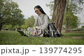 A hand-held shot of a young beautiful female student in casual clothes, wearing glasses and headphones, sitting on the grass under a tree in a city park and using a laptop while typing on the keyboard 139774295