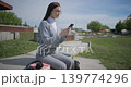 A hand-held shot of a young female student in casual clothes wearing glasses sitting on a concrete bench in an urban park on a sunny summer day using a mobile phone. 139774296
