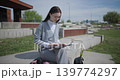 The shot is handheld and shows a young female student in casual clothes wearing glasses sitting on a concrete bench in an urban park and working on a tablet. 139774297