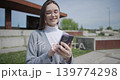 A hand-held shot of a young female student in casual clothes wearing glasses sitting on a concrete bench in an urban park on a sunny summer day using a mobile phone. 139774298