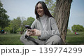 The shot is handheld and shows a young female student in casual clothes wearing glasses sitting on the grass under a tree in a park and using a mobile phone. 139774299