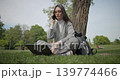 A hand-held shot of a young, beautiful female student in casual clothes, wearing glasses and headphones, sitting on the grass under a tree in a city park, using a laptop and talking on a mobile phone. 139774466
