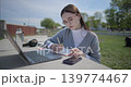 A hand-held shot of a young female student in casual clothes and glasses using a laptop and taking notes with a pen in a notebook in an urban park. 139774467